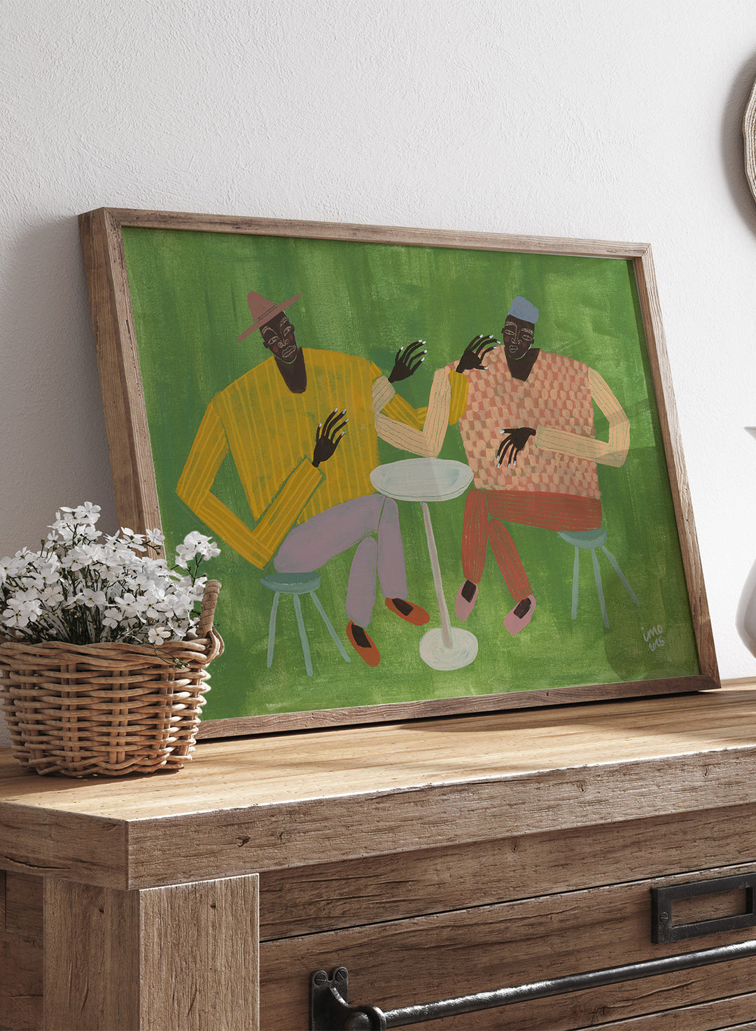 Framed artwork of two people sitting at a table on a green background. In Good Company by Studio IMO (Ibi Meier-Oruitemeka) is a modern figurative art print featuring two figures at a table in a bold contemporary wall poster design.