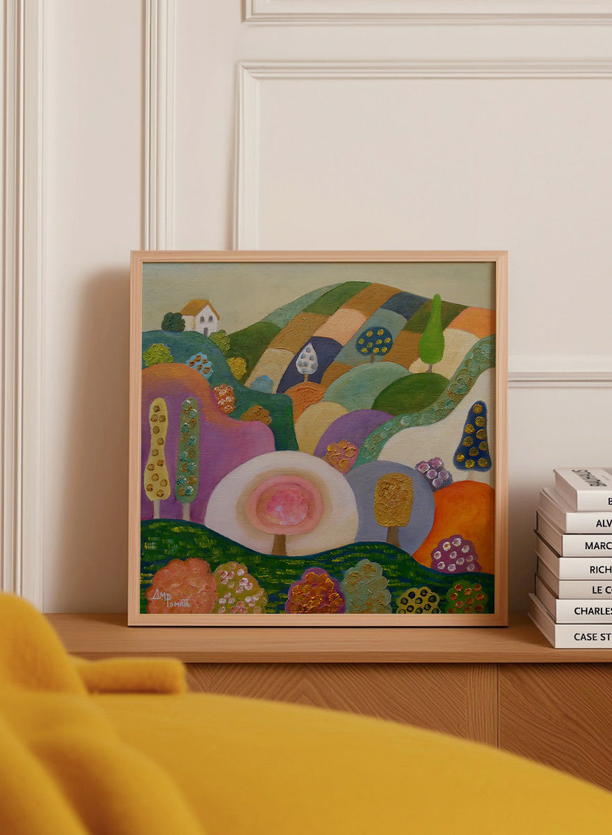 A colorful abstract landscape painting in a black frame, featuring stylized hills, trees, flowers, and a small house. The artwork has a playful, patchwork-like design with multiple vibrant colors including green, purple, orange, pink, blue, and yellow, set against a light beige wall. Shop Whimsical Hillside Village by Ángeles M. Pomata, a colorful landscape art print with rolling hills, patterned trees, and a dreamy village feel.