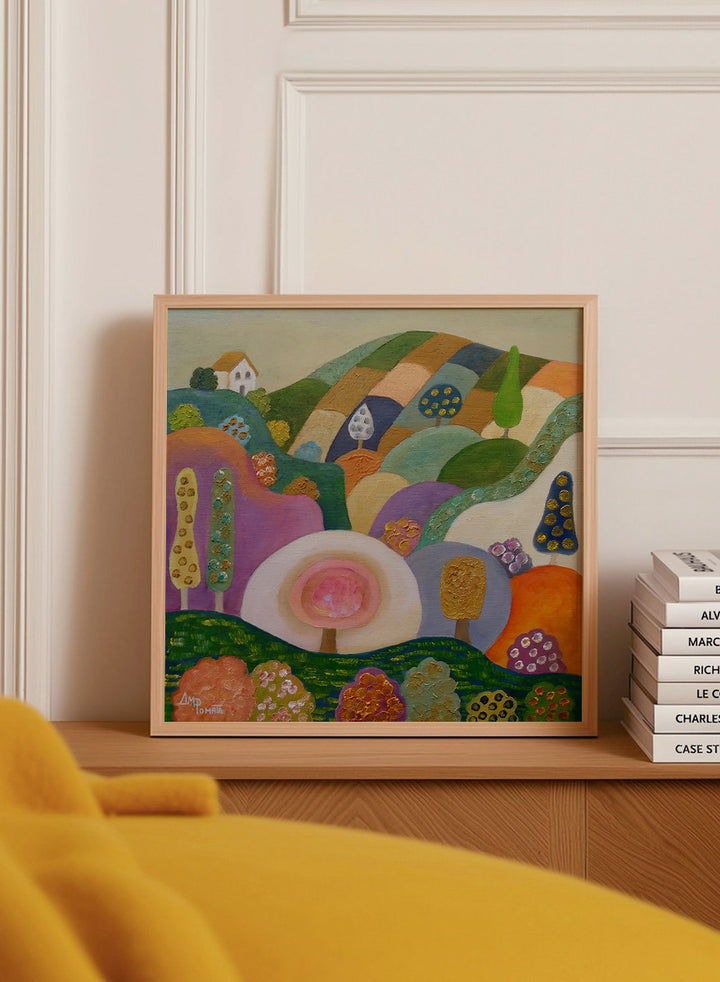 A colorful abstract landscape painting in a black frame, featuring stylized hills, trees, flowers, and a small house. The artwork has a playful, patchwork-like design with multiple vibrant colors including green, purple, orange, pink, blue, and yellow, set against a light beige wall. Shop Whimsical Hillside Village by Ángeles M. Pomata, a colorful landscape art print with rolling hills, patterned trees, and a dreamy village feel.