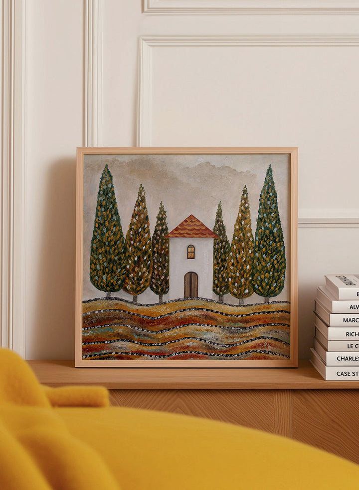 A framed landscape art print depicting a small white house with a red-tiled roof, surrounded by tall green cypress trees and wavy golden-brown hills. Shop Tuscan Cypress House by Angeles M. Pomata, an autumn landscape art print featuring a white country house, tall cypress trees, terracotta roof, and warm earthy tones.