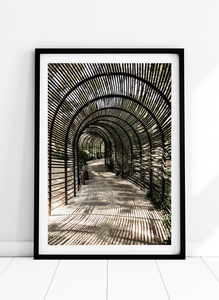 A framed art print featuring a perspective view of a garden pathway enclosed in a dark arched trellis structure, casting striped shadows on the ground. Shadow Garden by Shot by Clint architectural photography print featuring a curved wooden walkway, striped shadows, garden plants, and textured natural light.