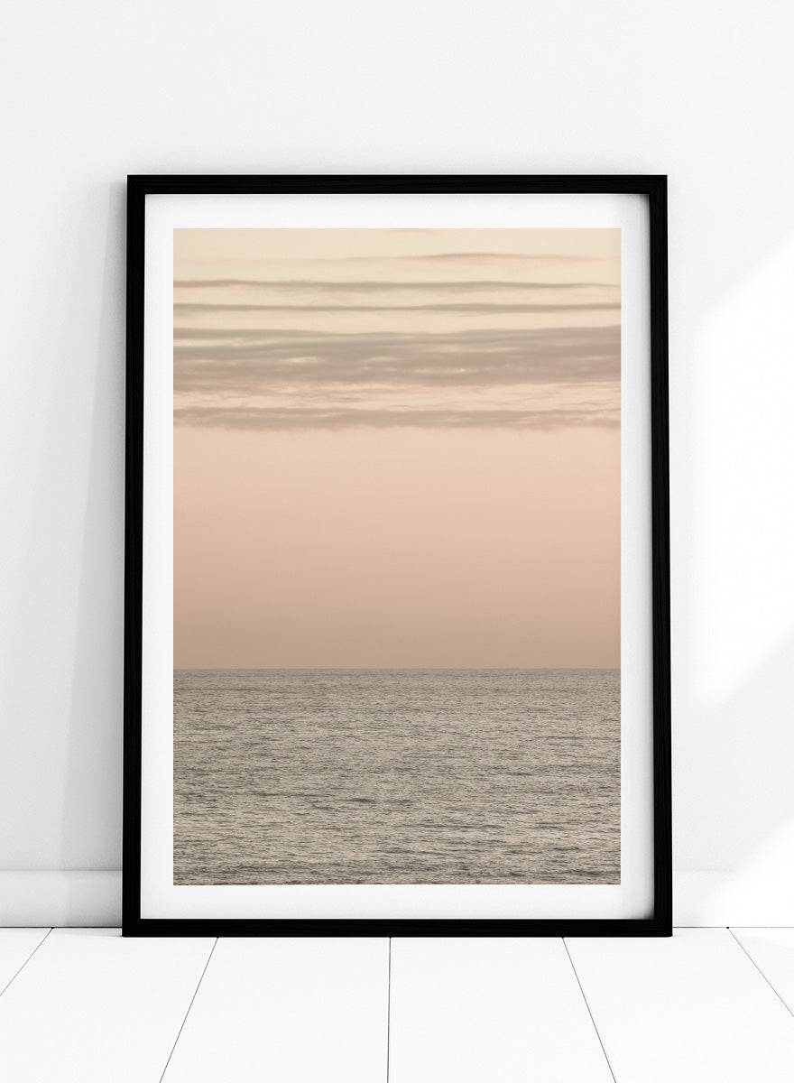 A minimalist framed art print depicting a calm seascape at dusk or dawn, with a soft peach and beige sky meeting a muted gray-blue ocean horizon. Serene Horizons by Shot by Clint features a soft peach sky and calm ocean horizon in a minimal coastal photograph with airy neutral tones.