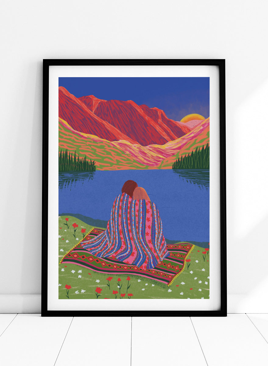 A framed art print featuring a stylized landscape illustration of two people sitting together on a patterned blanket by a blue lake, with red mountains and a yellow sun in the background. Cozy Lake Sunset art print by Ceyda Alasar features a lakeside embrace, vivid mountains, and a glowing sunset in rich, story-filled color.