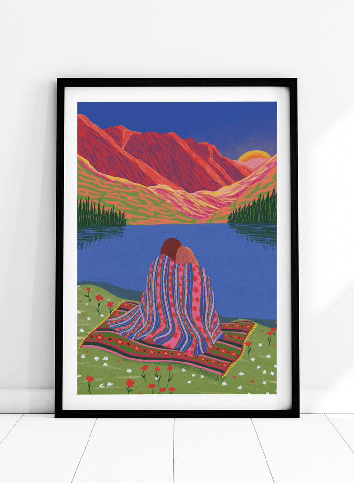 A framed art print featuring a stylized landscape illustration of two people sitting together on a patterned blanket by a blue lake, with red mountains and a yellow sun in the background. Cozy Lake Sunset art print by Ceyda Alasar features a lakeside embrace, vivid mountains, and a glowing sunset in rich, story-filled color.