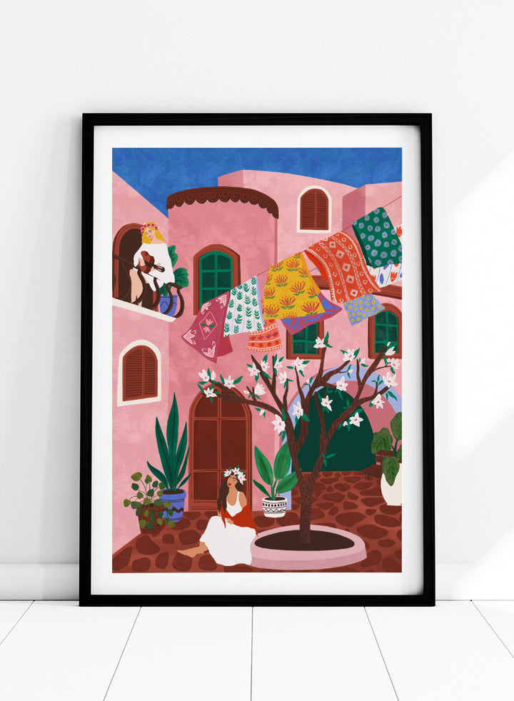 A framed art print featuring a colorful Mediterranean-style illustration of a pink courtyard with a woman sitting under a flowering tree, laundry hanging in the background, and a blue sky above. Pink Village Courtyard by Ceyda Alasar art print featuring pink architecture, patterned rugs, flowering trees, potted plants, and relaxed courtyard life.