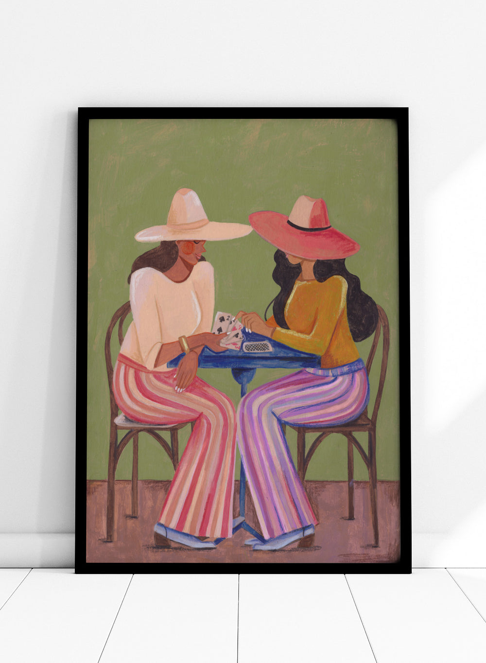 Two women playing cards at a table with colorful clothing and hats against a green background. Stylish Women Play Cards by Caroline Bonne Müller is a fine art print and wall poster featuring two fashionable women playing cards at a table.