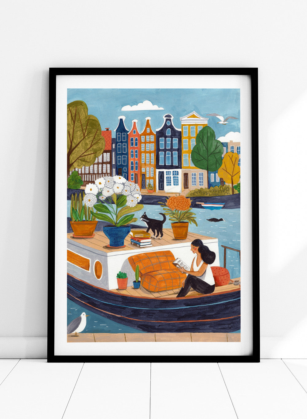 Illustration of a woman on a boat with flowers and a cat, surrounded by colorful houses. Cozy Canal Reading by Caroline Bonne Müller is a fine art print and wall poster featuring a woman reading on a canal boat with colorful European buildings in the background.