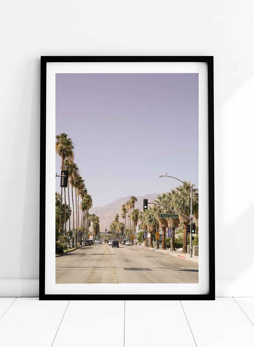 A framed art print featuring a scenic photograph of a palm-lined street in Palm Springs with mountains in the background. Palm-Lined Avenue by Shot by Clint travel photography print featuring a California road, tall palm trees, desert mountains, and soft sun-faded tones.