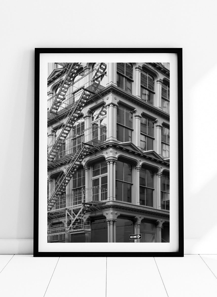 Black and white architectural photograph of a corner building facade with ornate windows and external fire escapes. Urban Geometry by Shot by Clint black-and-white architectural photography print featuring fire escapes, tall windows, ornate columns, and layered city detail.