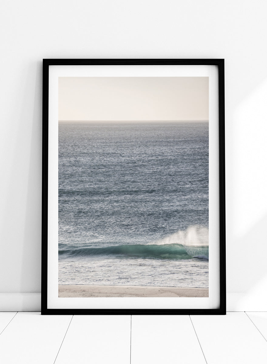 A framed photographic art print of an ocean scene, featuring a gentle wave breaking on a sandy beach under a pale sky. Tranquil Ocean Scene by Shot by Clint ocean photography print with a rolling wave, soft sea spray, and airy blue coastal tones.