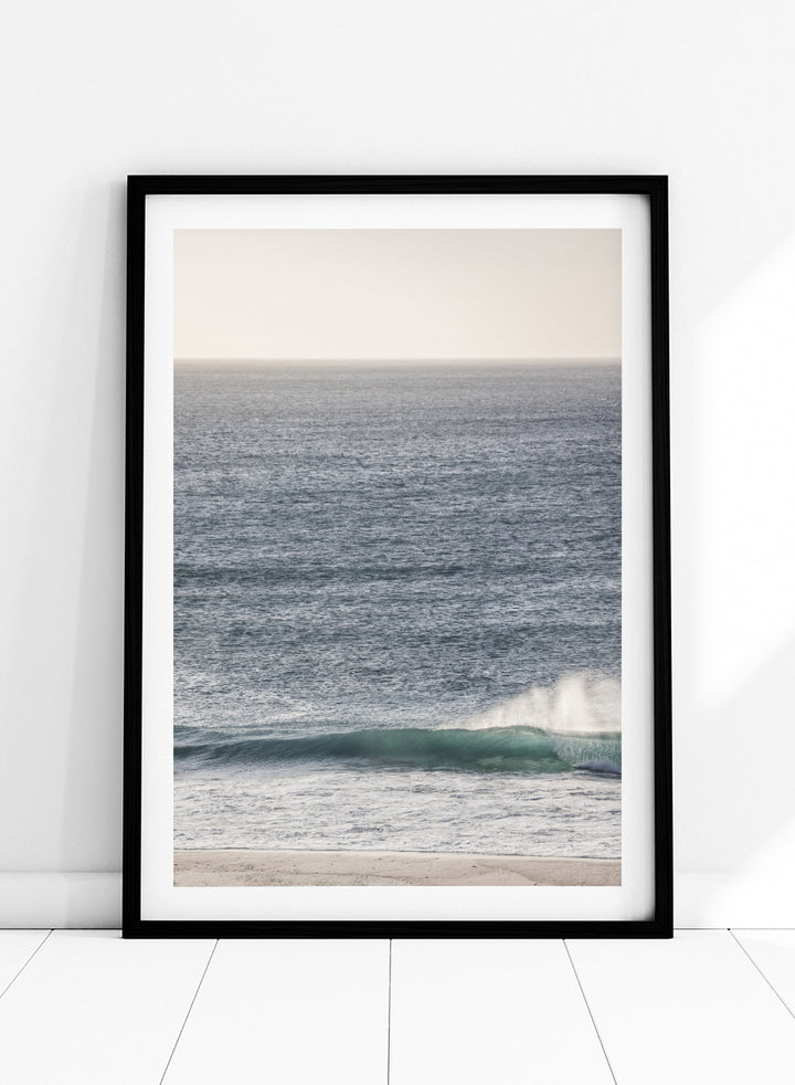 A framed photographic art print of an ocean scene, featuring a gentle wave breaking on a sandy beach under a pale sky. Tranquil Ocean Scene by Shot by Clint ocean photography print with a rolling wave, soft sea spray, and airy blue coastal tones.