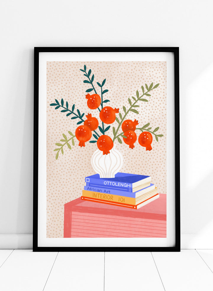 A framed art print featuring a stylized illustration of pomegranate branches in a white vase resting on a stack of books, set against a light beige dotted background. Cozy Tabletop Pomegranates by Bea Müller still life art print featuring red fruit branches, a sculptural vase, design books, and colorful interior styling.