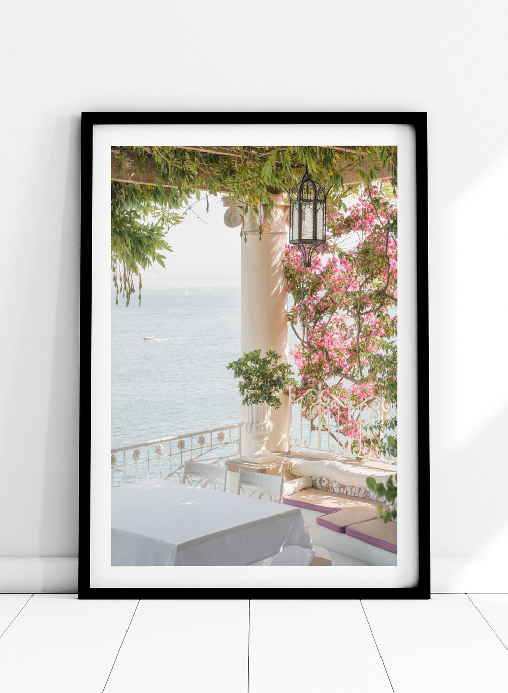 Outdoor seating area with flowers and ocean view. Pink Flowers Ocean View by Henrike Schenk is a coastal photography print featuring blooming flowers, Mediterranean architecture, ocean views, and soft natural light.