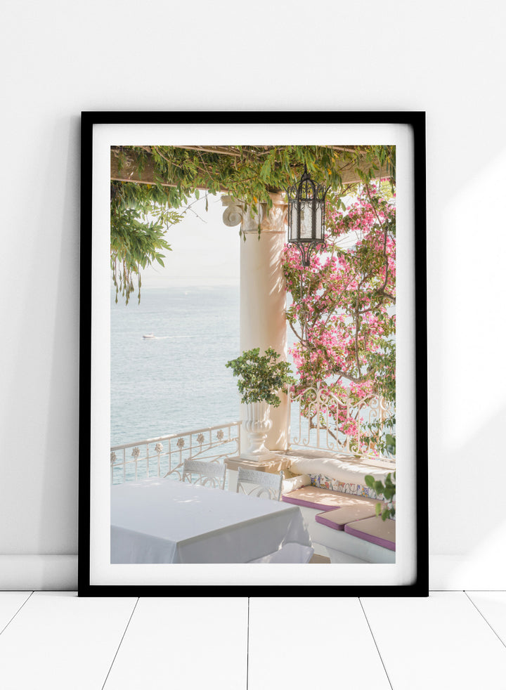 Outdoor seating area with flowers and ocean view. Pink Flowers Ocean View by Henrike Schenk is a coastal photography print featuring blooming flowers, Mediterranean architecture, ocean views, and soft natural light.