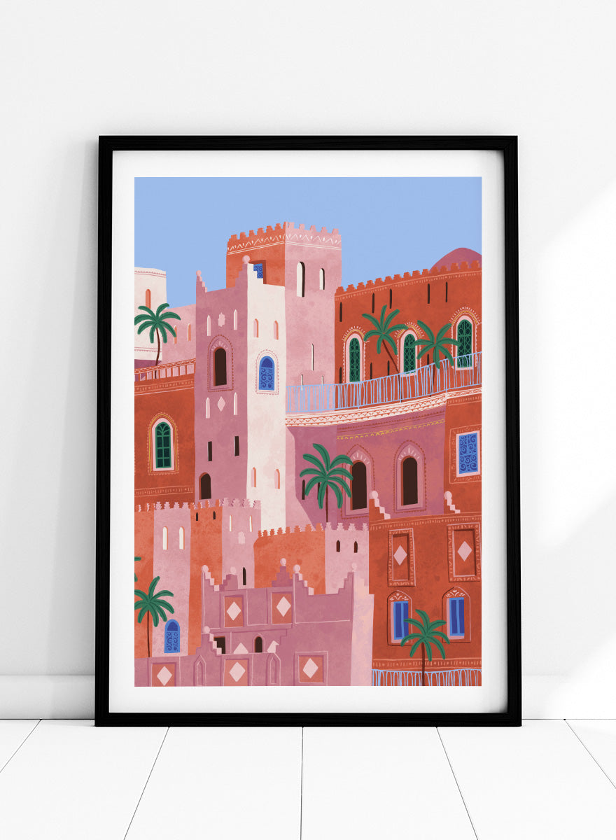 A framed art print featuring a stylized illustration of Moroccan-style buildings in shades of terracotta, pink, and cream, accented with green palm trees and blue window details, set against a light blue sky. Vibrant Moroccan City by Ceyda Alasar travel art print featuring layered terracotta architecture, arched windows, palms, and colorful Moroccan details.