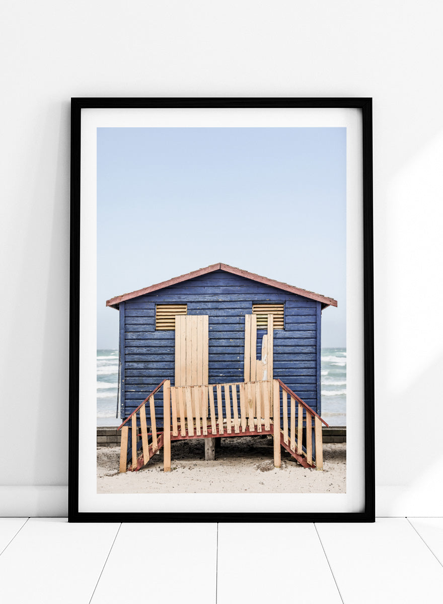 A framed photographic print of a blue wooden beach lifeguard hut on a sandy shore with the ocean and pale sky in the background. Beachside Retreat by Shot by Clint features a weathered blue beach hut and soft ocean backdrop in a calm coastal photography print.