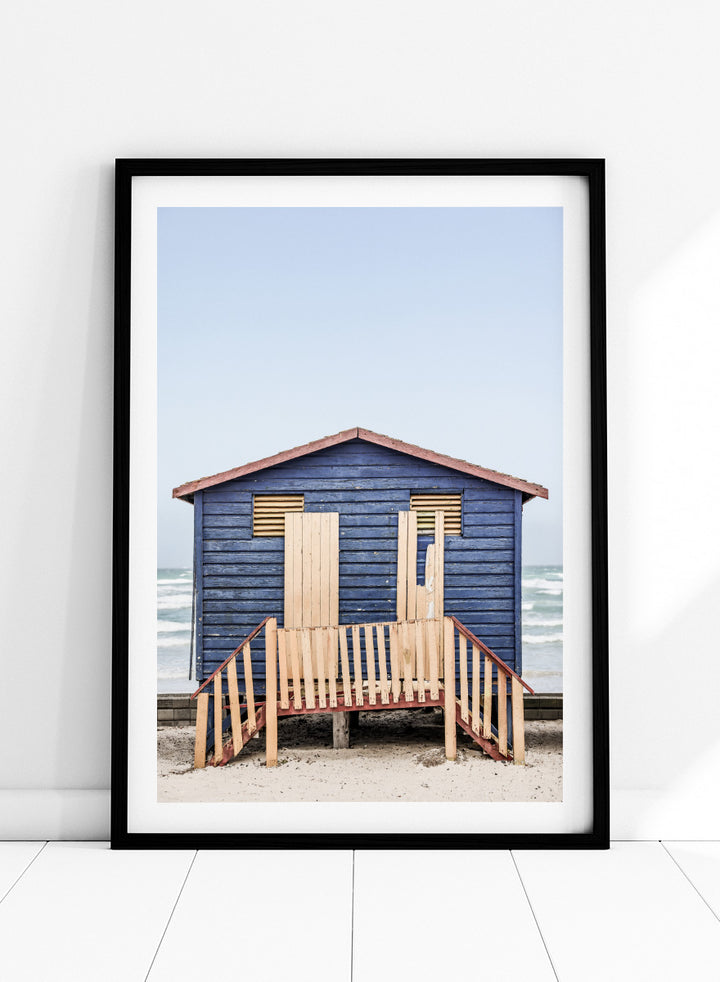A framed photographic print of a blue wooden beach lifeguard hut on a sandy shore with the ocean and pale sky in the background. Beachside Retreat by Shot by Clint features a weathered blue beach hut and soft ocean backdrop in a calm coastal photography print.