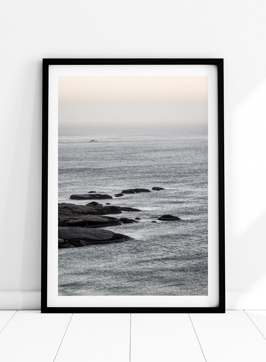 A framed black and white coastal landscape photograph featuring a calm sea, distant rocks, and a pale sky. Silver Sands by Shot by Clint coastal photography print featuring silver-blue water, dark shoreline rocks, and a soft misty horizon.