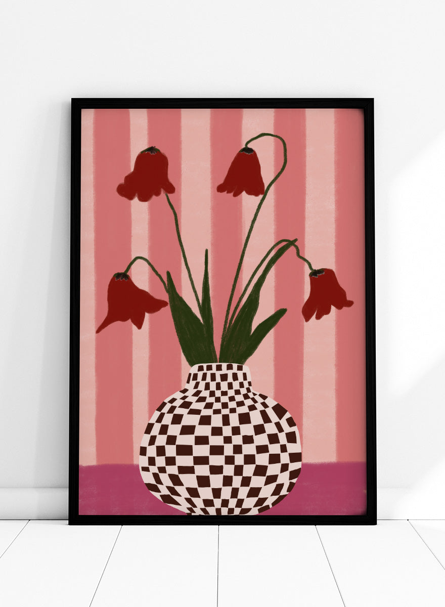 A stylized floral artwork showing four dark red bell-shaped flowers with long green stems rising from a black-and-white checkerboard vase, set against pink vertical stripes and a pink base. Whimsical Flowers Checkered Vase by The Art Concept still life art print featuring red flowers, a checkered vase, and a soft pink striped background.