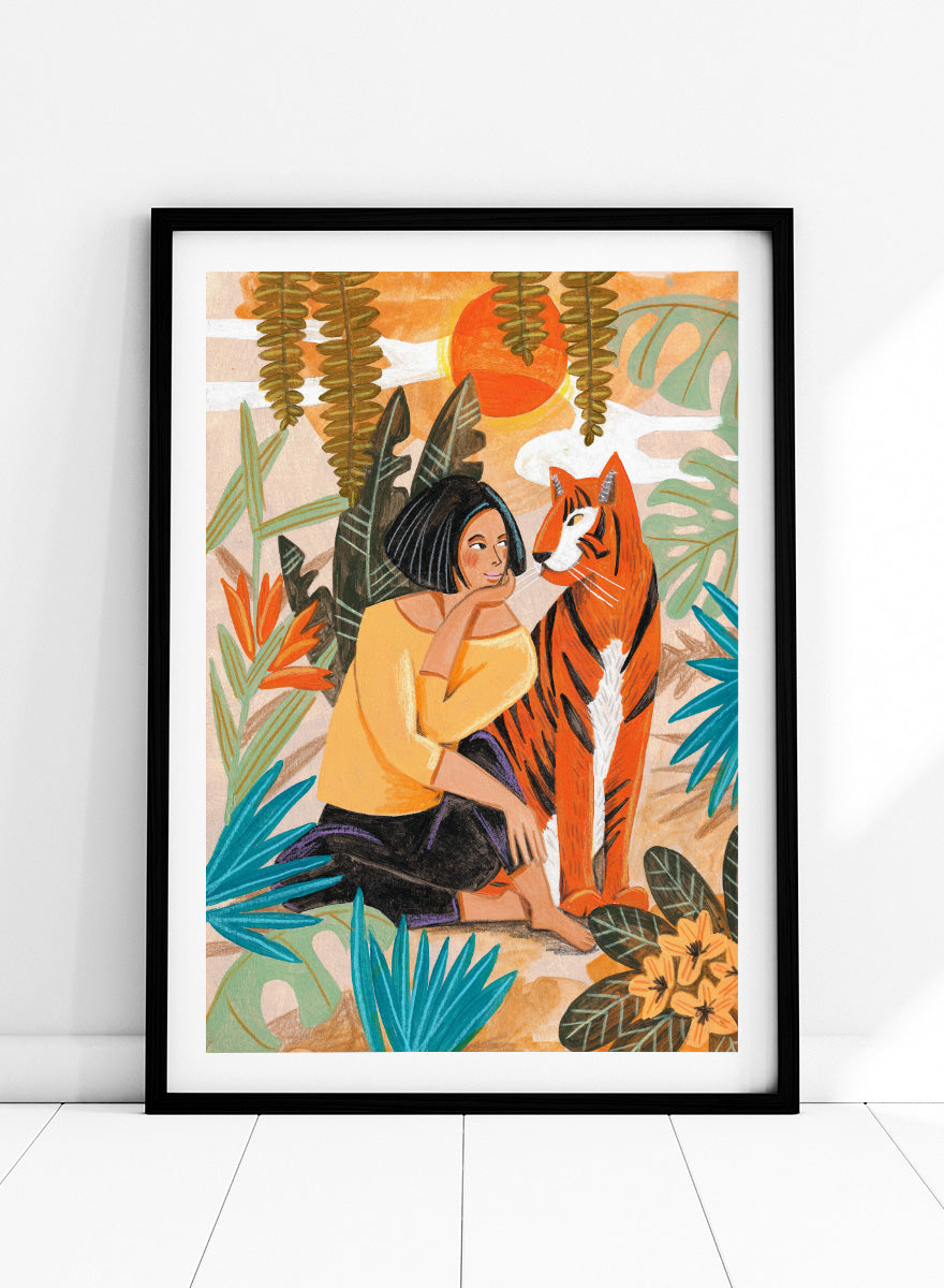 A framed art print featuring a stylized illustration of a woman in a yellow top and black skirt sitting beside an orange tiger, surrounded by tropical leaves and a warm orange sky. Woman with Tiger by Caroline Bonne Müller tropical art print featuring a woman beside a tiger among lush foliage, bold color, and hand-drawn texture.