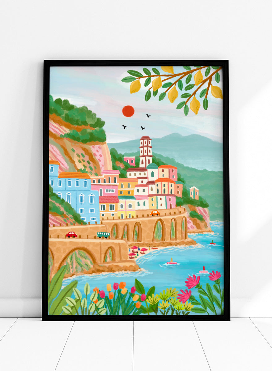 A colorful illustrated coastal town scene with pastel buildings, a stone bridge, blue water, mountains, flowers, and hanging yellow leaves. Shop Colorful Amalfi Coast by Nidhi Kachhadiya, a vibrant Italian coastal wall art print inspired by the Amalfi Coast, pastel villages, blue sea, and lemon trees.