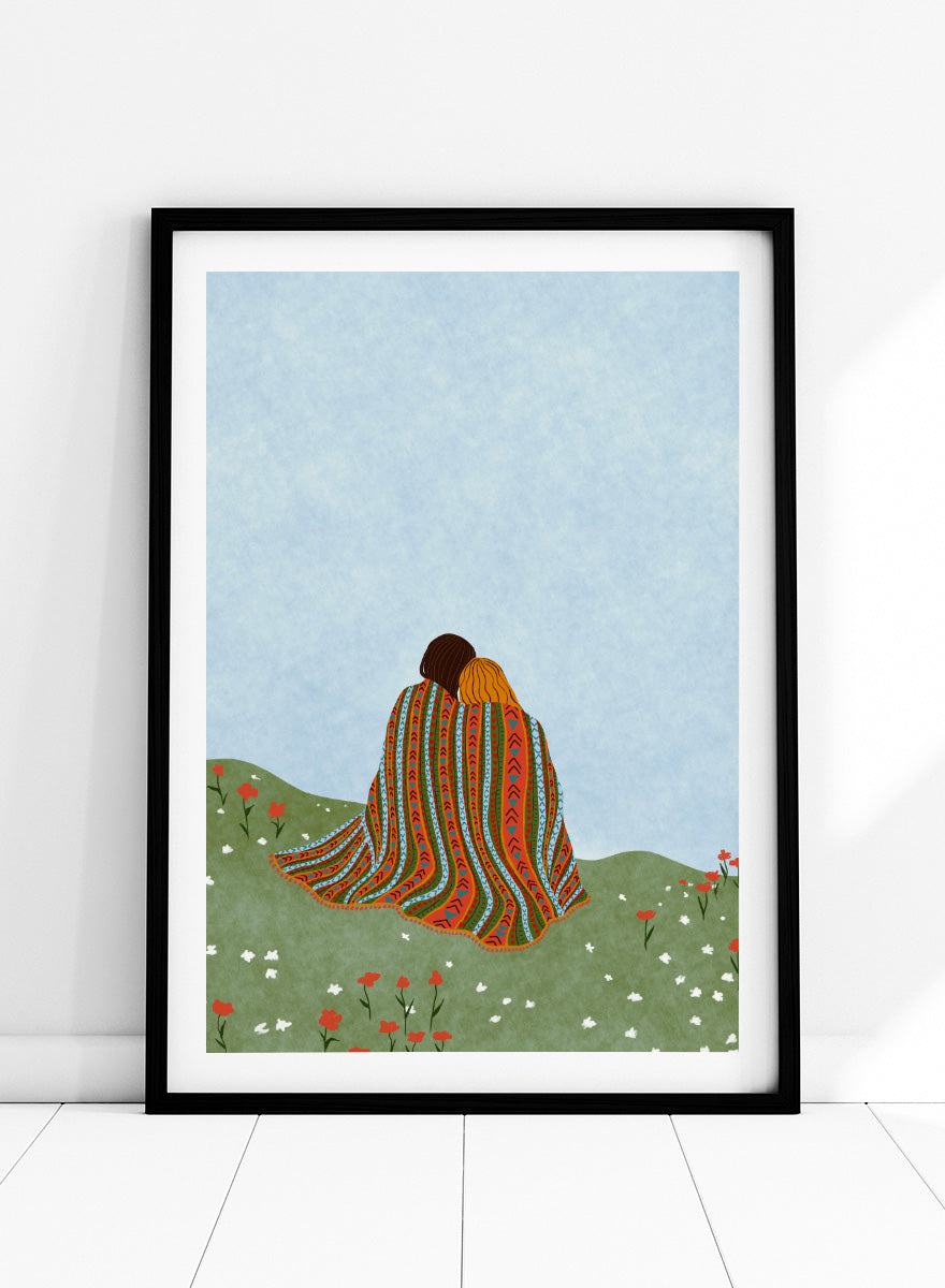 A framed art print featuring an illustration of two people sitting on a grassy hill, wrapped in a vibrant, patterned blanket under a light blue sky. Cozy Hillside Embrace by Ceyda Alasar art print featuring two people wrapped in a patterned blanket among green hills and wildflowers.