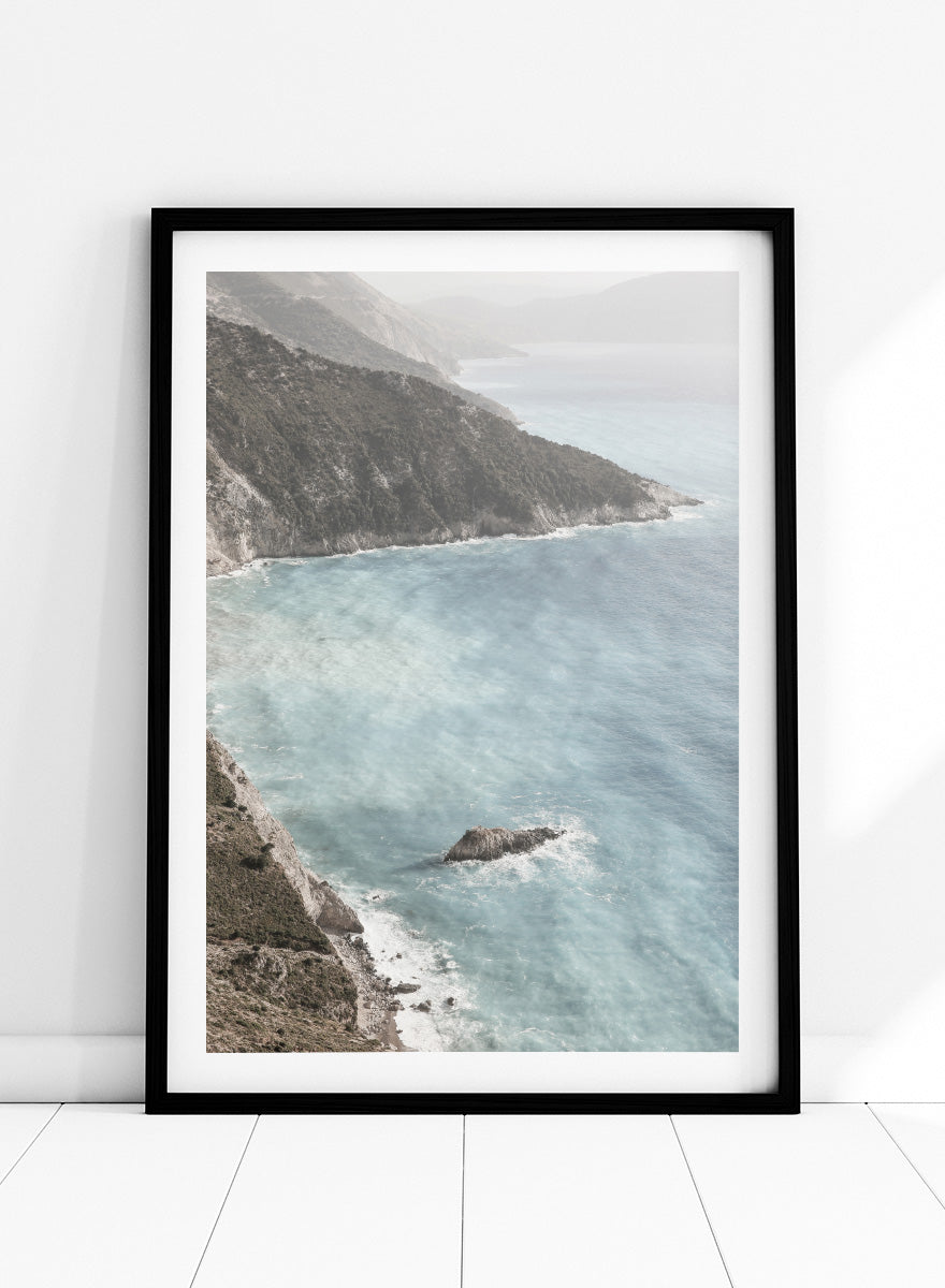 A framed photographic art print depicting a coastal landscape with a steep rocky cliff overlooking a calm, pale blue sea with a small rock formation in the foreground. Coastal Serenity by Shot by Clint coastal photography print featuring pale blue water, rugged cliffs, rocky shores, and a soft hazy horizon.
