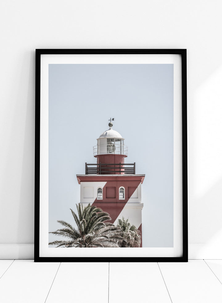 A framed art print featuring a red and white lighthouse with palm trees at the bottom, set against a pale sky background. Beacon of Solitude by Shot by Clint lighthouse photography print featuring red-and-white architecture, palm trees, arched windows, and a pale coastal sky.