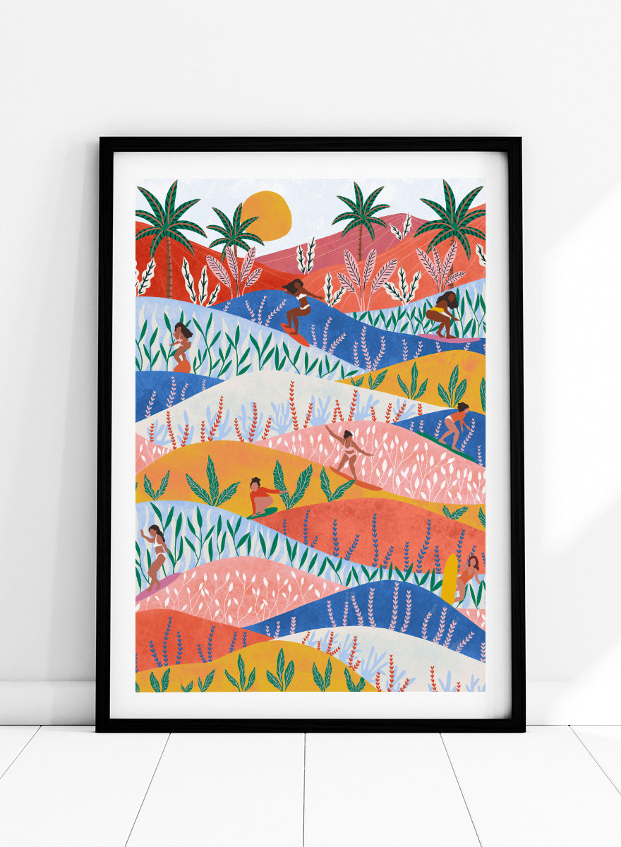 A framed art print featuring a colorful, stylized landscape illustration with rolling hills, palm trees, a sun, and small human figures in a folk-art style. Tropical Surf Girls by Ceyda Alasar surf art print featuring women riding waves through a colorful palm-filled landscape in bold tropical tones.