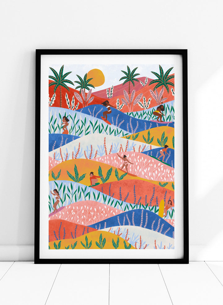 A framed art print featuring a colorful, stylized landscape illustration with rolling hills, palm trees, a sun, and small human figures in a folk-art style. Tropical Surf Girls by Ceyda Alasar surf art print featuring women riding waves through a colorful palm-filled landscape in bold tropical tones.