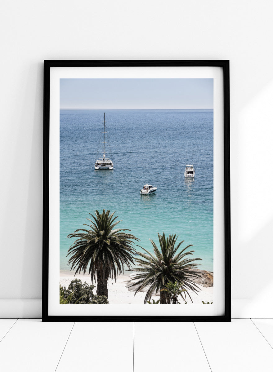 A framed coastal photograph showing a calm blue ocean with three boats, turquoise water near a white sandy shore, and two palm trees in the foreground. Tropical Serenity by Shot by Clint coastal photography print featuring turquoise water, sailboats, palm trees, and a bright tropical shoreline.