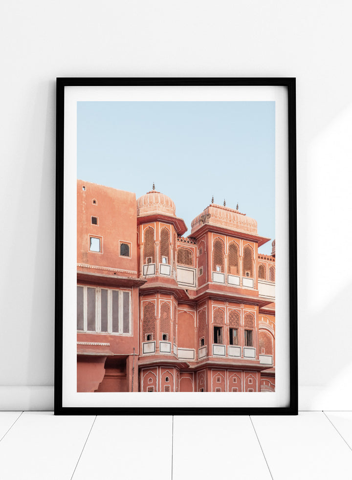 A framed art print featuring a photograph of the iconic pink sandstone Rajasthani palace, Udaipur, with white architectural details under a clear light blue sky. Terracotta Splendor by Shot by Clint architectural photography print featuring ornate palace windows, domed balconies, carved details, and warm coral tones.