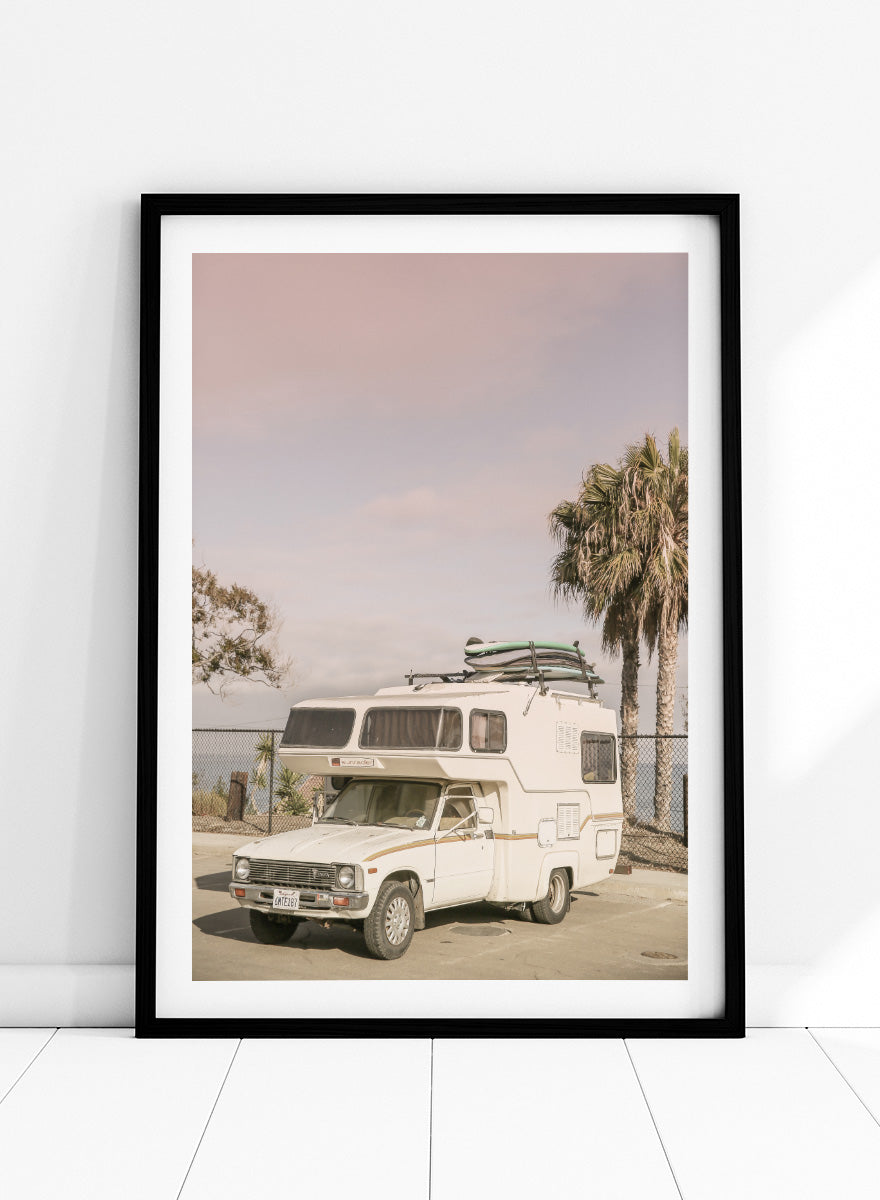 A vintage white camper truck parked outdoors near palm trees and a fence, with a surfboard strapped to its roof rack. Vintage Camper Parked by Shot by Clint travel photography print featuring a retro camper, surfboards, palm trees, and sun-faded coastal tones.