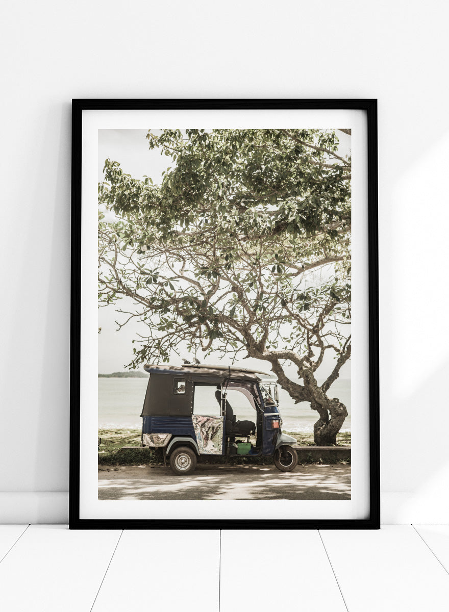 A framed photographic print featuring a small blue and white auto rickshaw parked under a large leafy tree beside a body of water. Tuk Tuk Tour by Shot by Clint travel photography print featuring a blue tuk tuk, surfboard, coastal tree, and relaxed seaside setting.