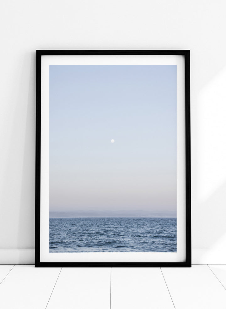A minimalist framed art print depicting a calm blue ocean under a pale sky with a small visible moon. Moonlit Horizon by Shot by Clint coastal photography print featuring a pale moon, distant shoreline, open blue water, and soft lavender evening tones.