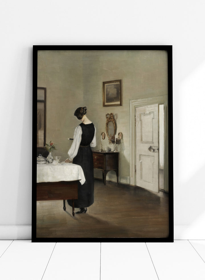 Woman in a black dress standing in a room with a table and mirror. In the Dining Room by Carl Holsøe is a vintage Danish interior art print featuring a woman in a softly lit dining room with muted tones and classic Scandinavian style.