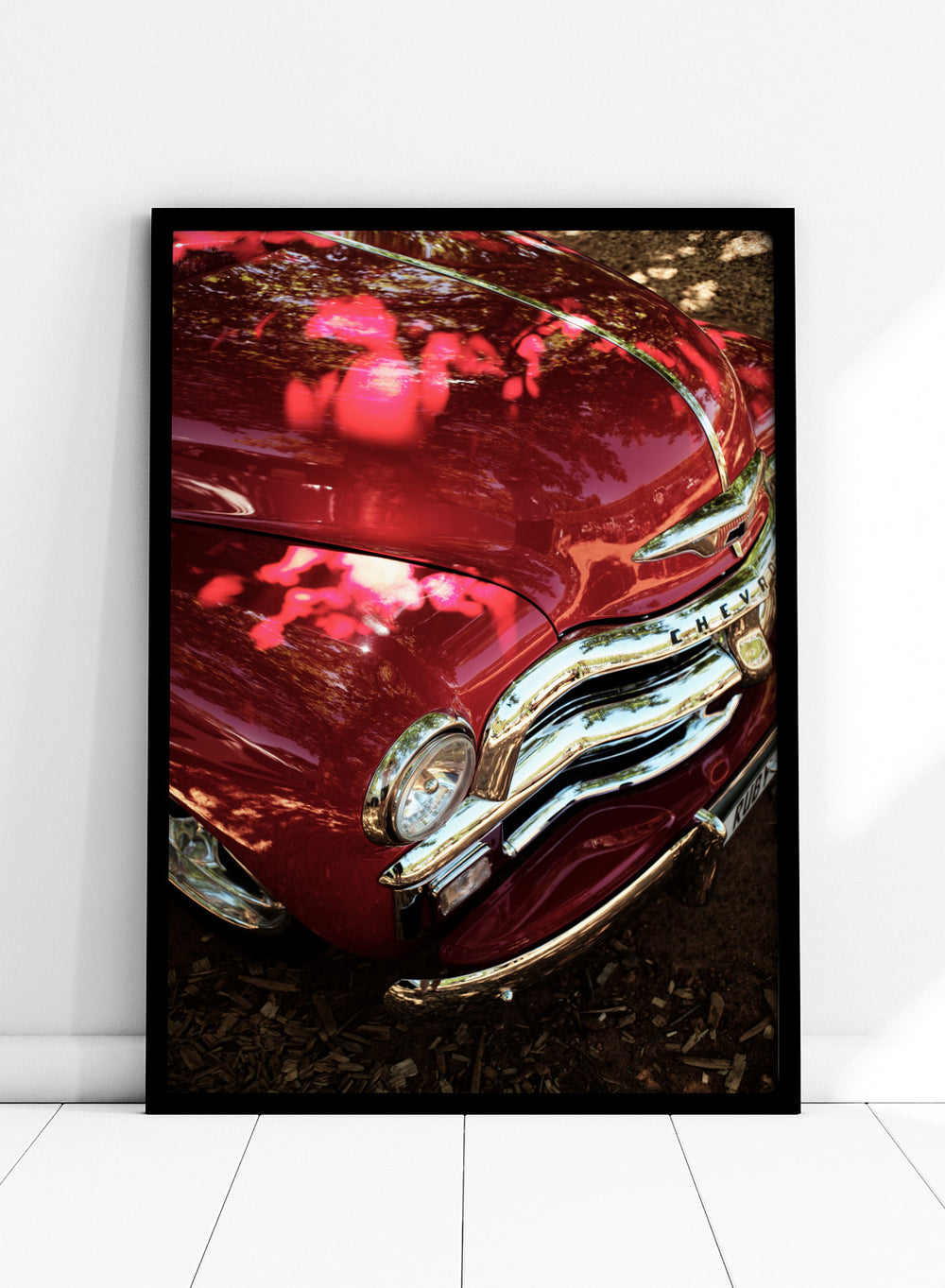 Close-up of a vintage red Chevrolet car body with chrome grille and reflections of trees on the glossy paint, photographed outdoors on mulch. Vintage Red Car by Shot by Clint automotive photography print featuring a classic Chevrolet, deep red paint, polished chrome, and sculptural vintage details.