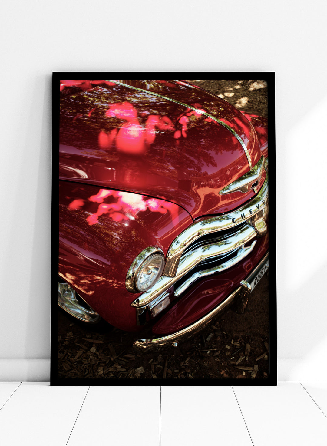 Close-up of a vintage red Chevrolet car body with chrome grille and reflections of trees on the glossy paint, photographed outdoors on mulch. Vintage Red Car by Shot by Clint automotive photography print featuring a classic Chevrolet, deep red paint, polished chrome, and sculptural vintage details.