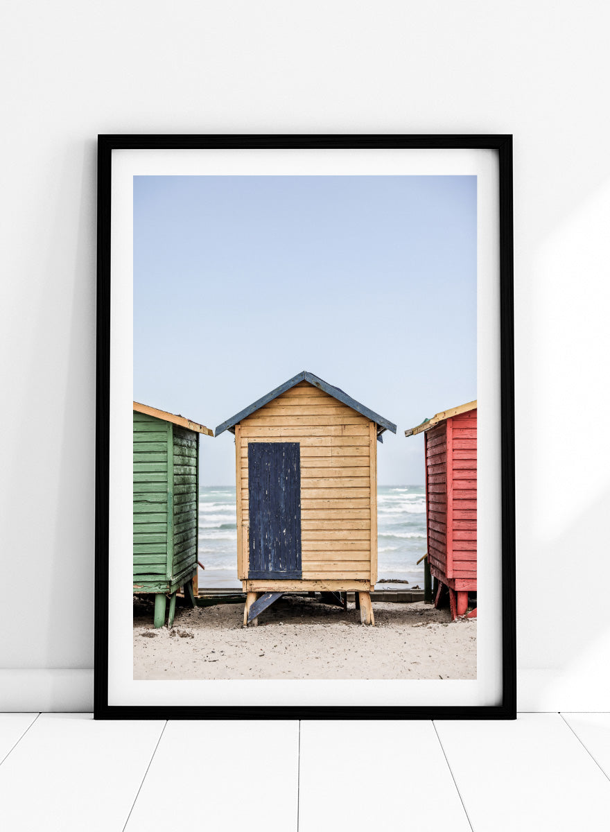 A framed photographic print of three colorful wooden beach huts on a sandy beach, with the central hut in natural wood and a dark blue door, flanked by green and red huts under a pale blue sky. Colorful Beach Huts by Shot by Clint coastal photography print featuring green, natural wood, and red huts with weathered texture and ocean views.