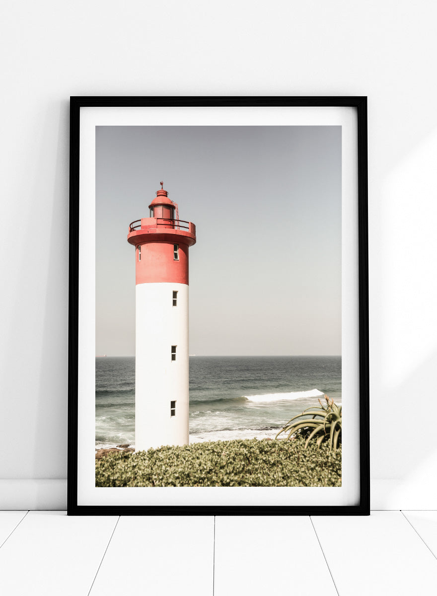 A framed photographic art print of a red and white lighthouse standing on a grassy shore with the ocean and sky in the background. Beacon by the Sea by Shot by Clint coastal photography print featuring a red-and-white lighthouse, ocean waves, pale sky, and coastal greenery.