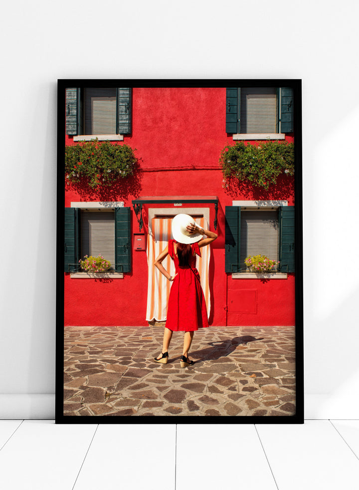 Woman in a red dress and white hat standing in front of a red wall with green shutters. Red Art Print by Anette Ohlendorf featuring a woman in a red dress standing against a red building. Officially licensed travel photography with strong color and symmetry.
