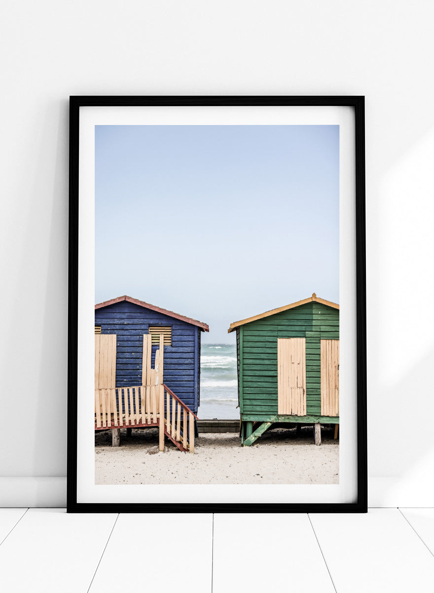 A framed photographic print of two wooden beach huts on a sandy beach under a pale blue sky, with one hut in dark blue and the other in green, both with light wood shutters and doors. Coastal Huts Harmony by Shot by Clint coastal photography print featuring blue and green beach huts, sandy ground, weathered wood, and ocean views.