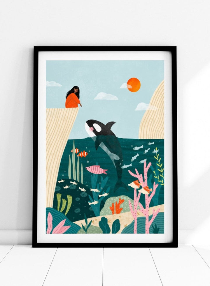 A framed art print featuring a whimsical underwater scene with an orca, coral, fish, and a woman in an orange dress standing on a cliff edge under a sun. Girl Watches Ocean Life by Bea Müller premium framed fine art print featuring a child watching an orca swim through a colorful underwater world.