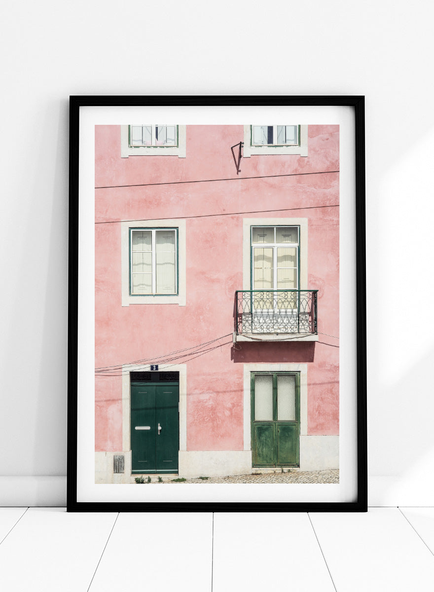 A framed art print featuring a photograph of a pink building facade with green doors, white-framed windows, and a small black metal balcony. Pink Façade Harmony by Shot by Clint architectural photography print featuring a pink exterior, green doors, white windows, and an iron balcony.