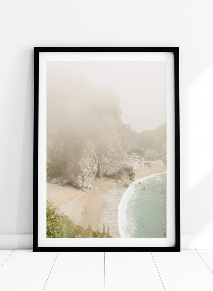 A framed photographic print of a misty coastal beach scene with a small waterfall, sandy shore, and pale blue-green ocean waves. Misty Coastal Haven by Shot by Clint coastal photography print featuring a secluded cove, rocky cliffs, pale turquoise water, and a soft veil of mist.