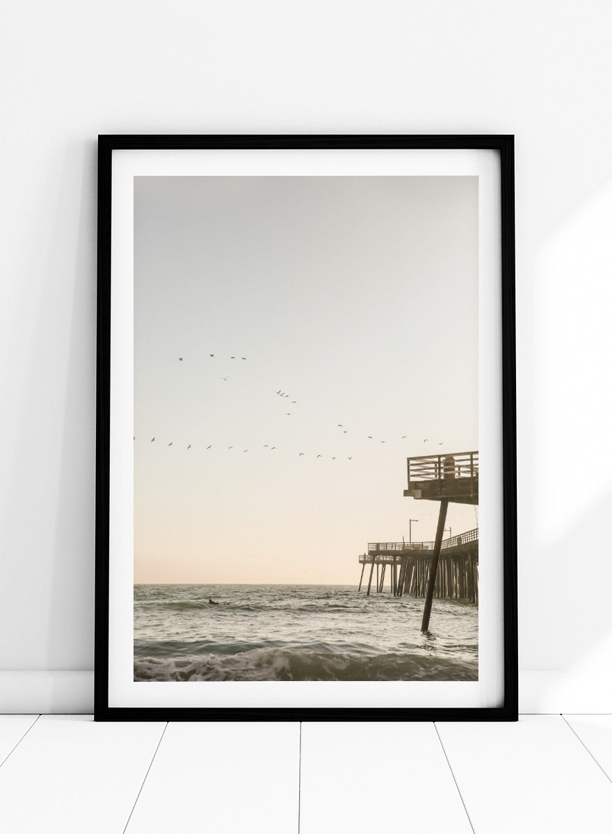 A framed photographic art print depicting a pier extending over a calm ocean under a pale sky, with birds flying across the scene. Seaside Serenity by Shot by Clint coastal photography print featuring ocean waves, a weathered pier, flying birds, and soft sunlit tones.