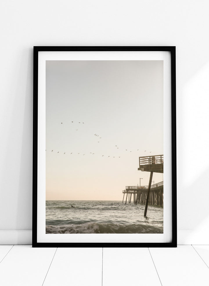 A framed photographic art print depicting a pier extending over a calm ocean under a pale sky, with birds flying across the scene. Seaside Serenity by Shot by Clint coastal photography print featuring ocean waves, a weathered pier, flying birds, and soft sunlit tones.