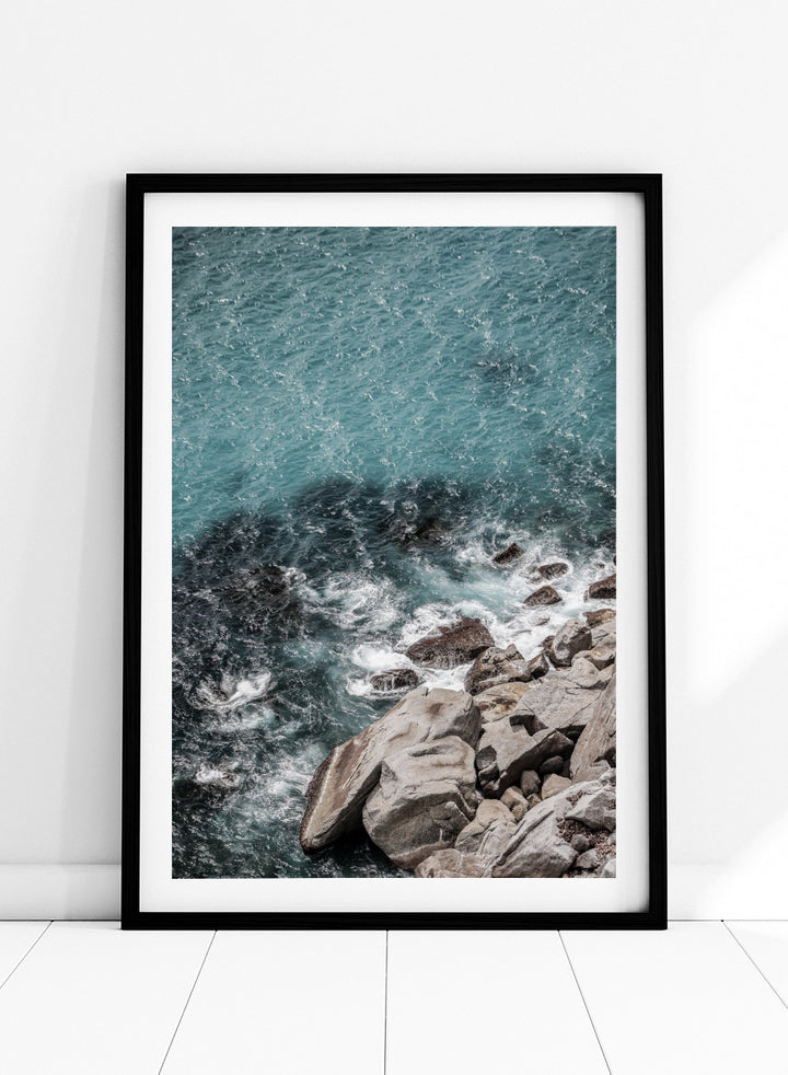A framed photographic art print depicting a top-down view of turquoise ocean water washing over dark rocks and a rocky shoreline. Ocean’s Edge Symphony by Shot by Clint aerial ocean photography print featuring turquoise water, white surf, coastal texture, and soft seafoam tones.