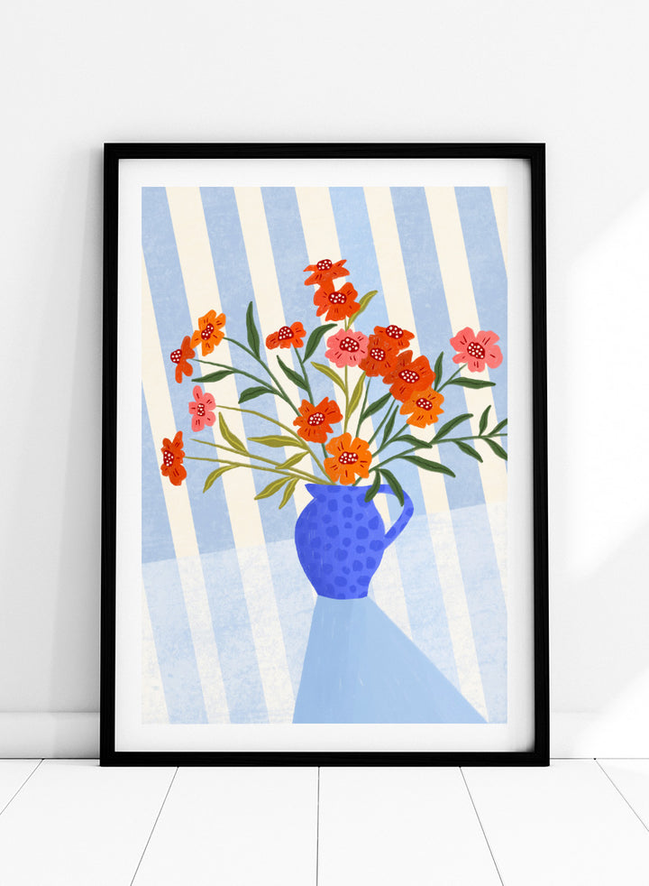 A framed art print featuring a stylized illustration of a blue patterned pitcher vase filled with orange, red, and pink flowers against a blue and white striped background. Cheerful Floral Pitcher by Bea Müller floral still life art print featuring orange and pink flowers in a bright blue jug with striped blue decor.