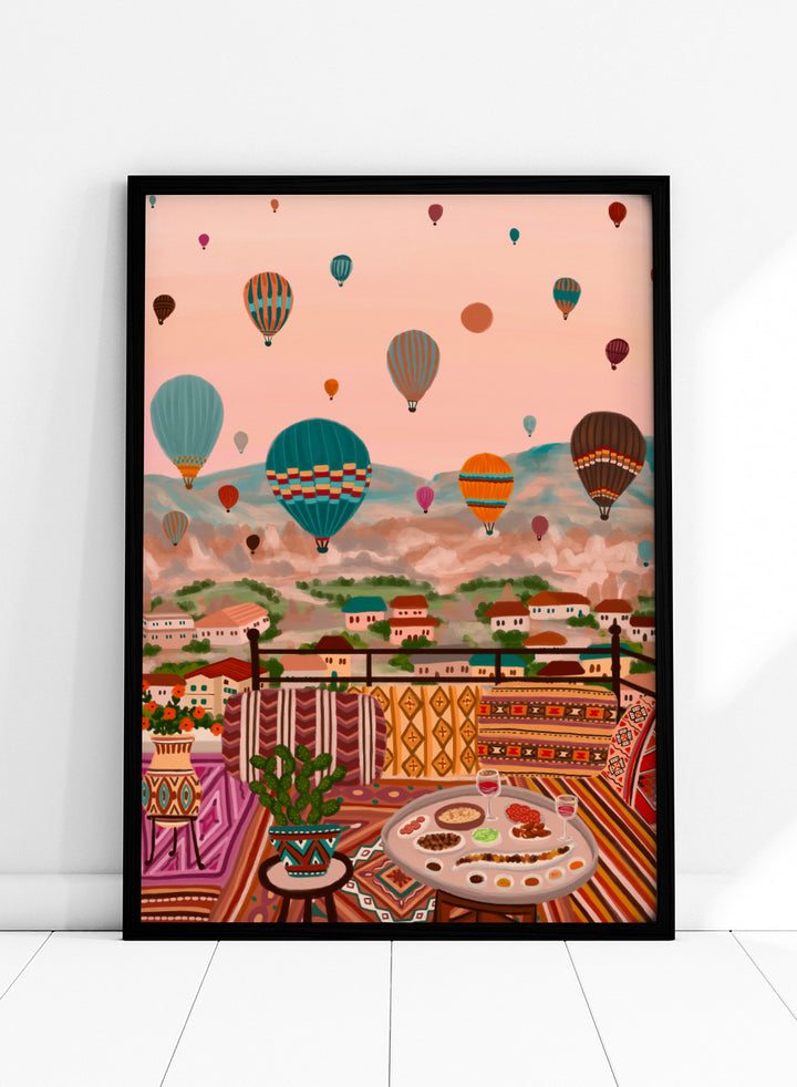 A colorful art print depicting a scenic view of a hillside town with mountains in the background, filled with hot air balloons. In the foreground, a patterned outdoor seating area features a table with food and a potted plant. Shop Balloons Over Rooftop Dinner by Nidhi Kachhadiya, a colorful travel wall art print featuring hot air balloons, rooftop dining, patterned textiles, and teal and coral tones.